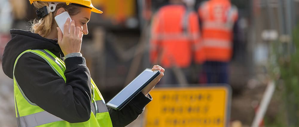 Worker wearing safety gear and accessing emergency response plans on a tablet with holding phone to ear
